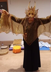Person im Drachenkostüm mit Maske und Flügelumhang in einem Seminarraum